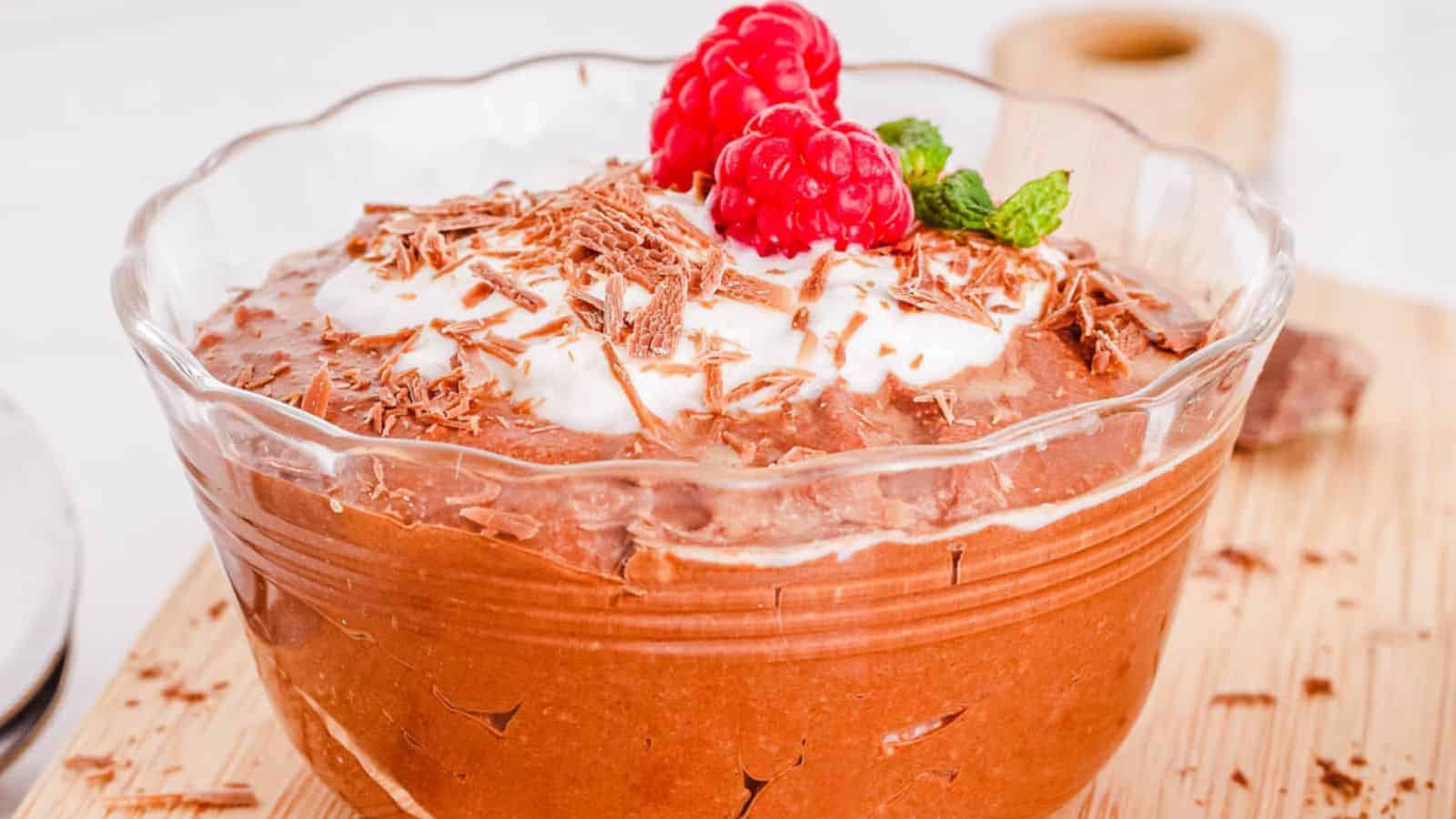 A glass bowl containing chocolate mousse is topped with whipped cream, chocolate shavings, two raspberries, and a mint sprig.