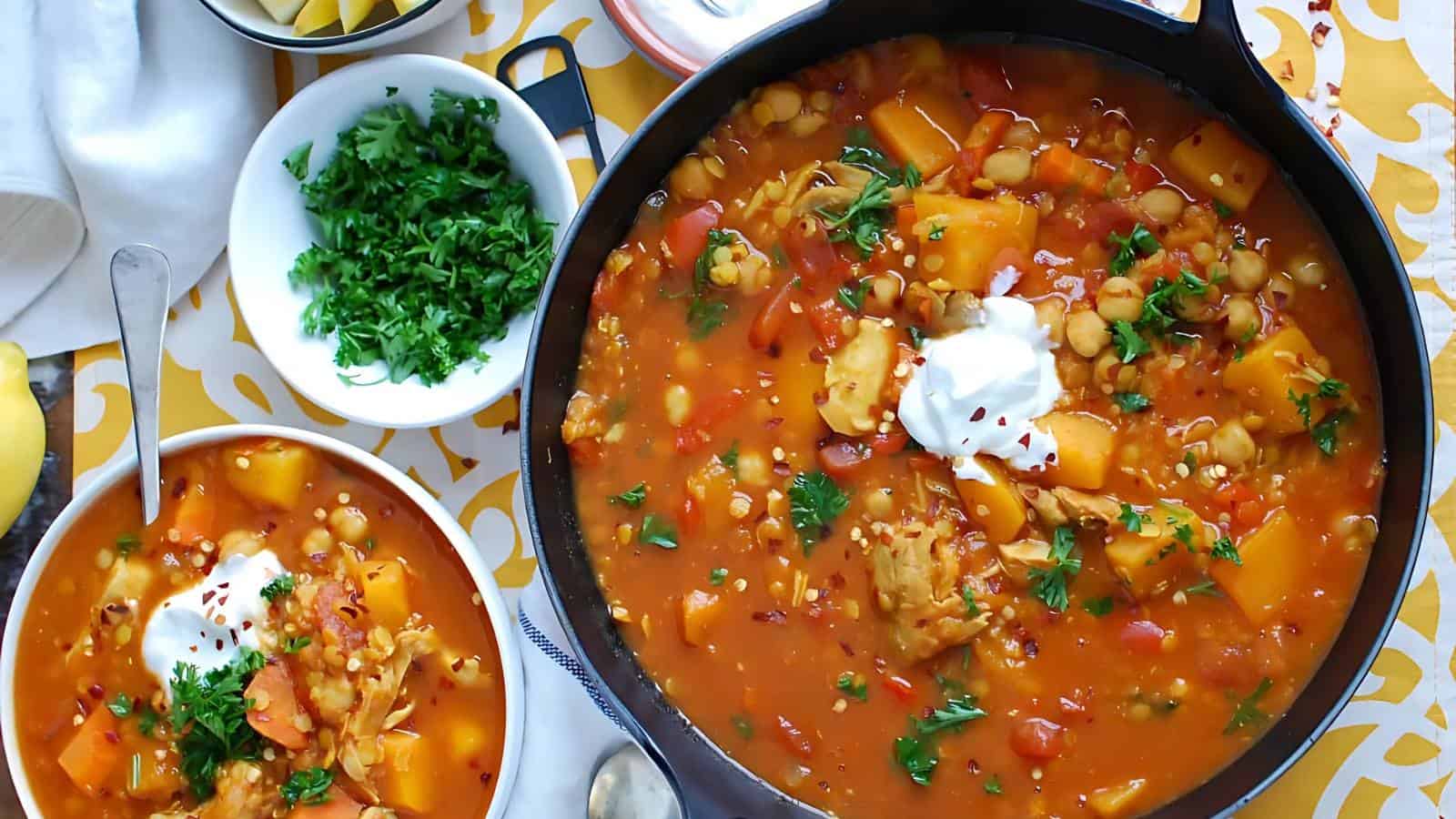 In the image, there is a pot and a bowl containing vegetable stew with chickpeas and herbs, topped with sour cream and red pepper flakes. A bowl of fresh chopped parsley is also present nearby, all on a patterned tablecloth.