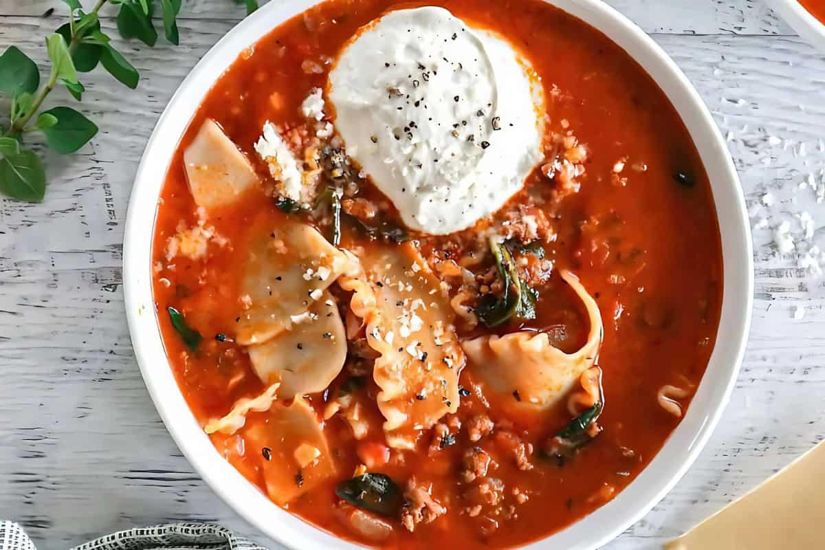 A bowl of lasagna soup topped with a dollop of creamy ricotta cheese and a sprinkle of black pepper. The soup features wide pasta noodles, crumbled sausage, and fresh spinach, all in a rich tomato broth. Fresh oregano is placed beside the bowl.
