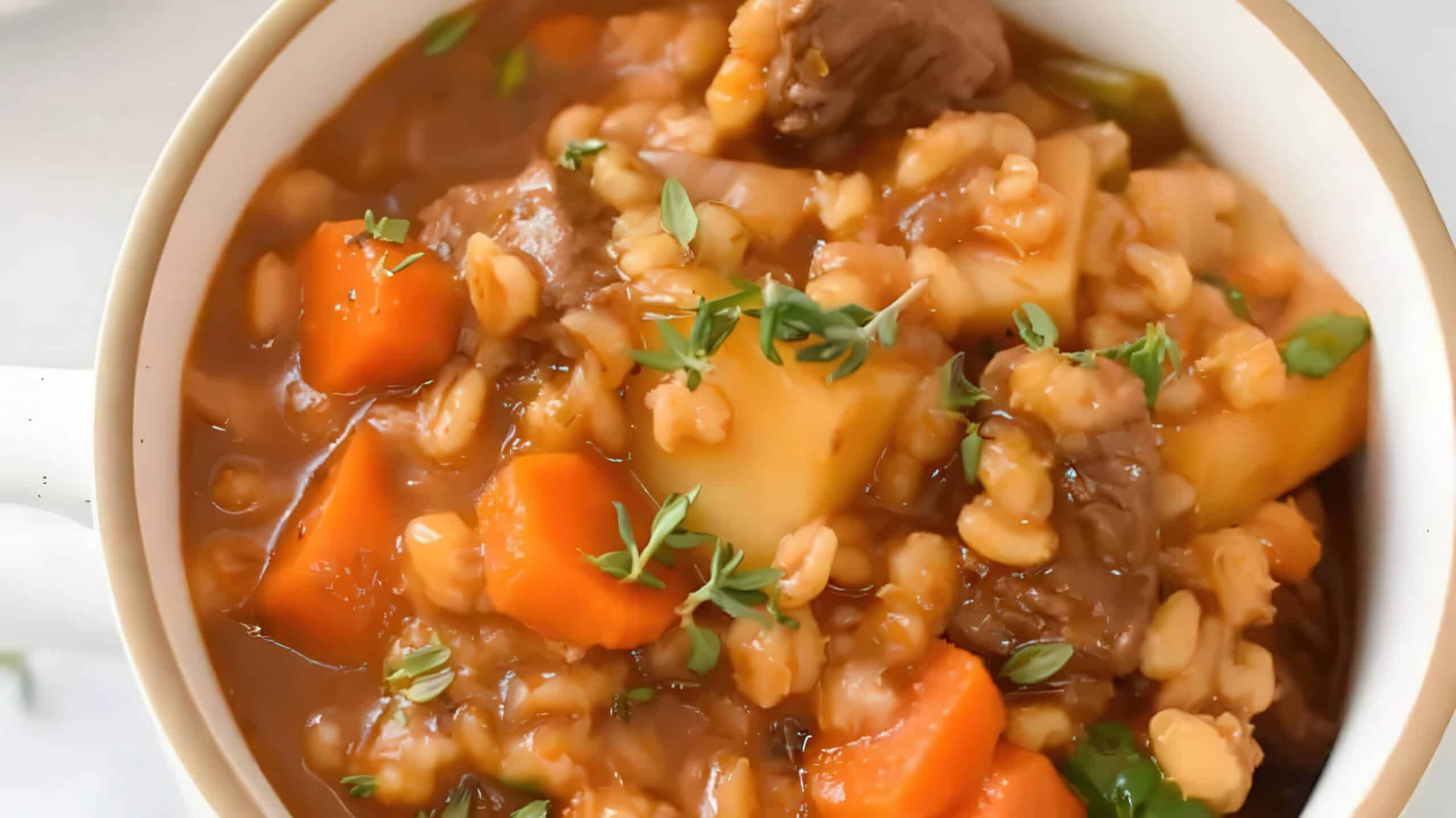 A close-up of a bowl of hearty beef stew with chunks of beef, potatoes, carrots, and barley, garnished with fresh thyme. The rich, savory broth is visible around the ingredients.