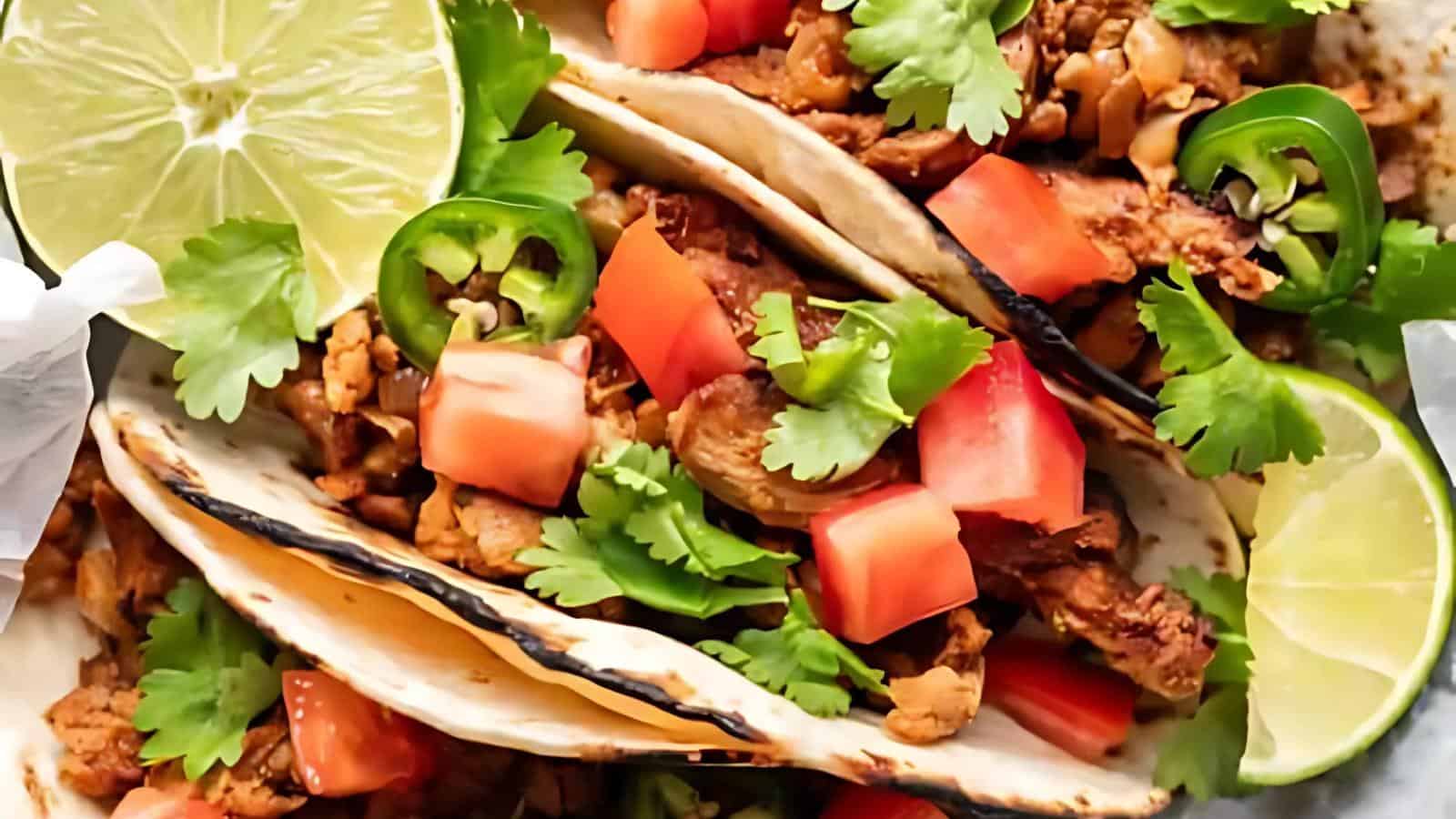 The image shows two tacos containing grilled meat, chopped tomatoes, sliced jalapeños, cilantro, and lime wedges.