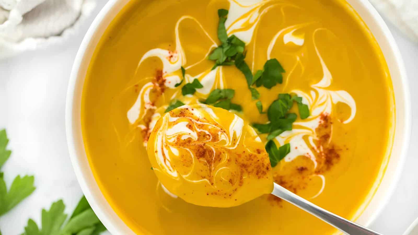 A bowl of creamy orange soup topped with swirls of cream, chopped parsley, and a sprinkle of paprika. A spoon is lifting a portion from the bowl.