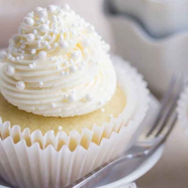 A vanilla cupcake with white frosting and pearl sprinkles sits on a white plate with a fork beside it.
