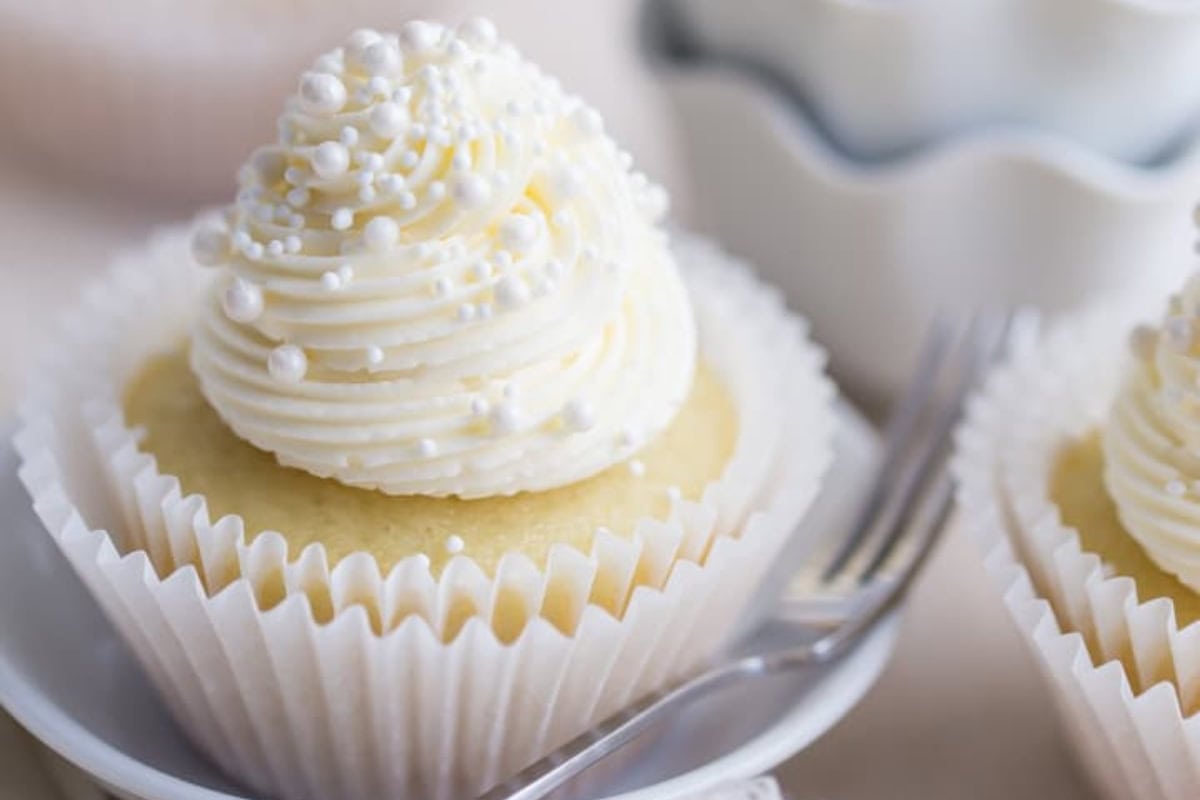 A vanilla cupcake with swirled white frosting topped with small white sugar pearls, placed in a white paper liner on a saucer. A fork rests beside it, and there are additional cupcakes and a fluted dish in the background.