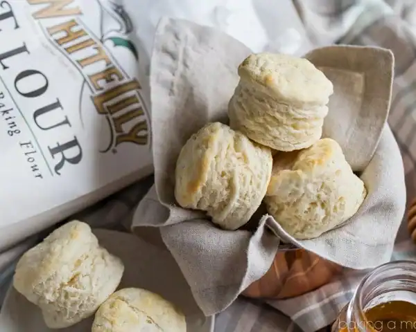 A basket of homemade biscuits on a cloth napkin, next to a bag of White Lily flour and a honey dipper.