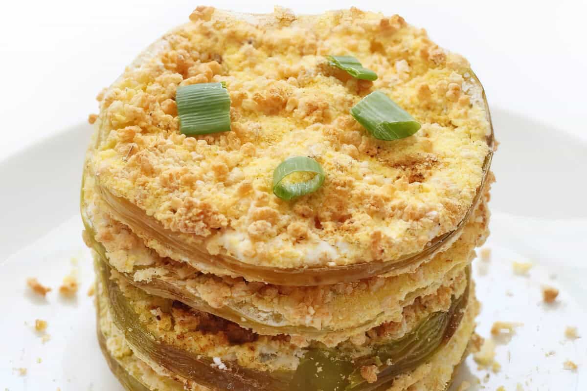 A white plate featuring a stack of breaded and fried tomato slices, topped with pieces of green onion.