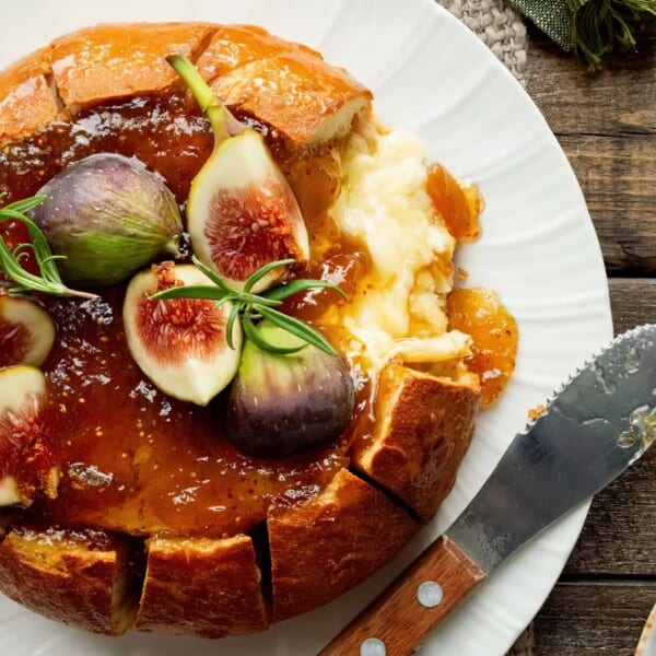 A round loaf of bread filled with melted cheese, topped with fig slices and rosemary, is served on a white plate with a spreading knife.