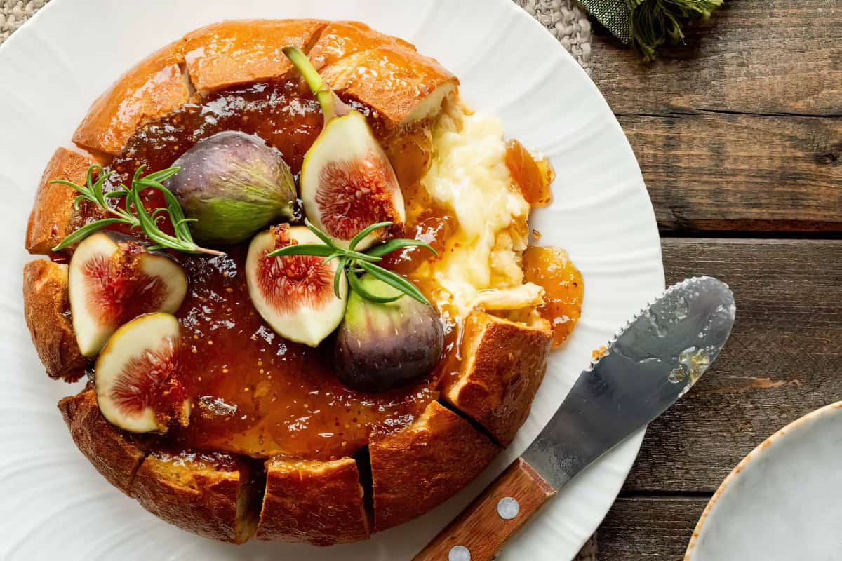 A round loaf of bread filled with melted cheese, topped with fig slices and rosemary, is served on a white plate with a spreading knife.