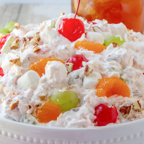 A bowl containing marshmallows, cherries, grapes, oranges, and pecans, with a cherry on top.