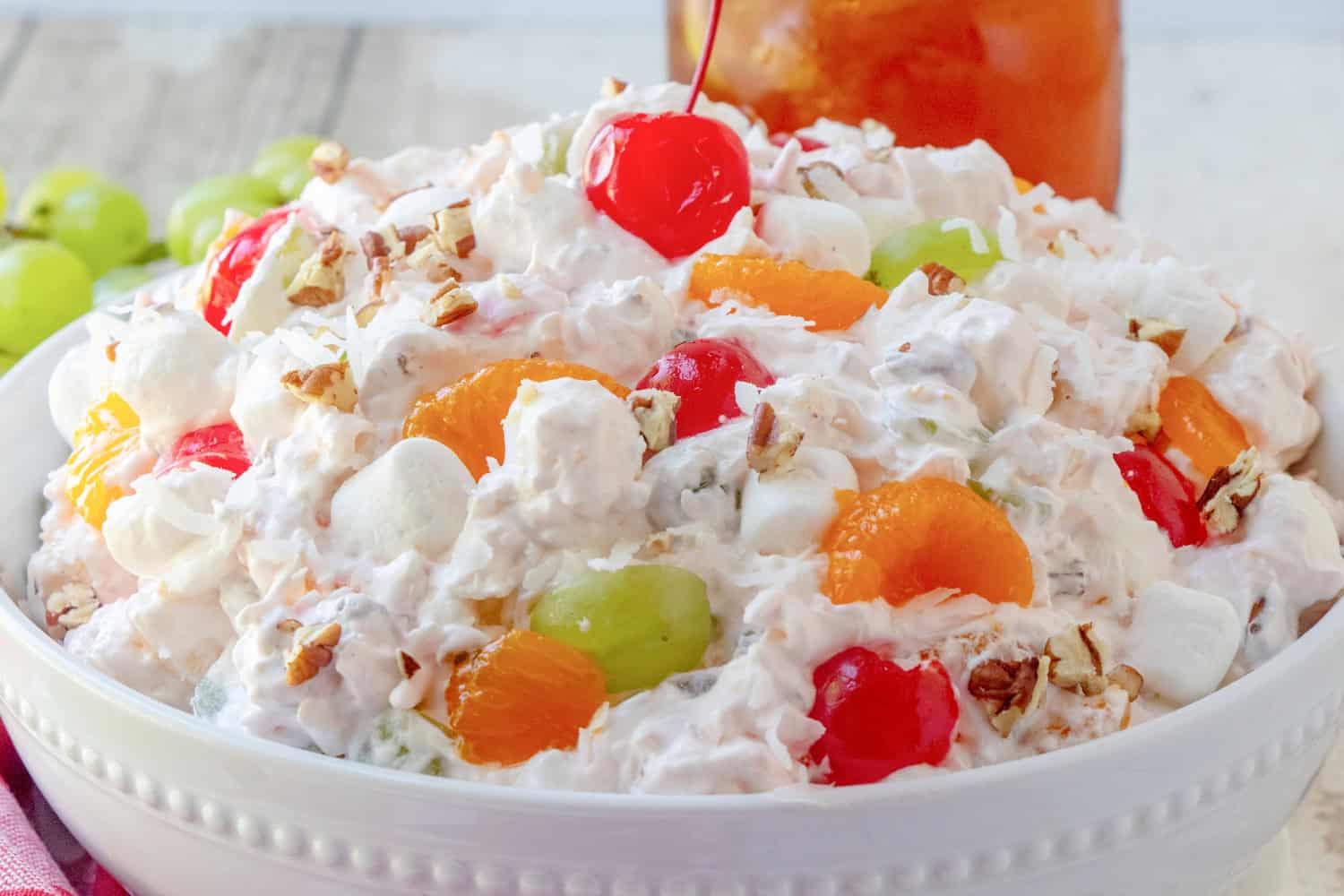 A bowl containing marshmallows, cherries, grapes, oranges, and pecans, with a cherry on top.