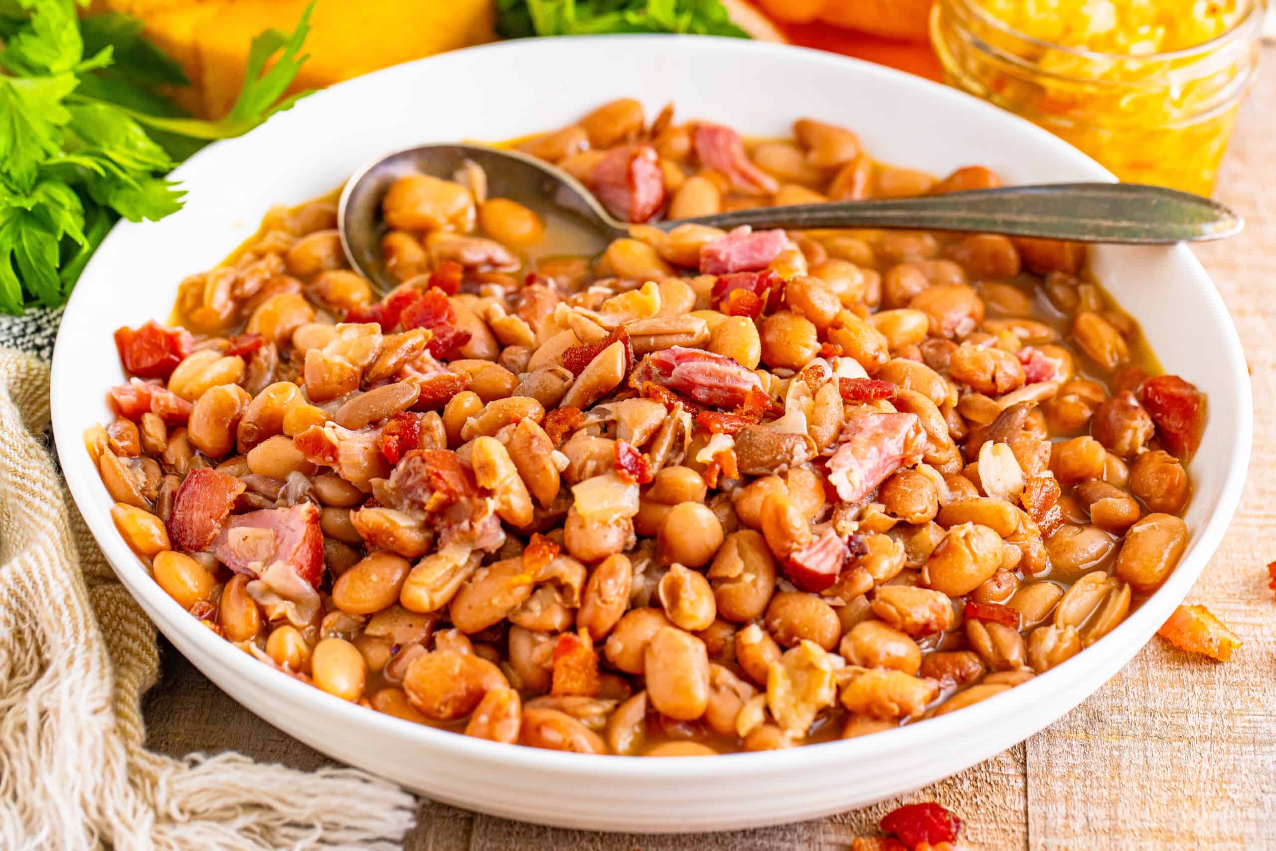 A bowl containing baked beans and pieces of bacon, with a spoon nearby, all on a wooden surface.