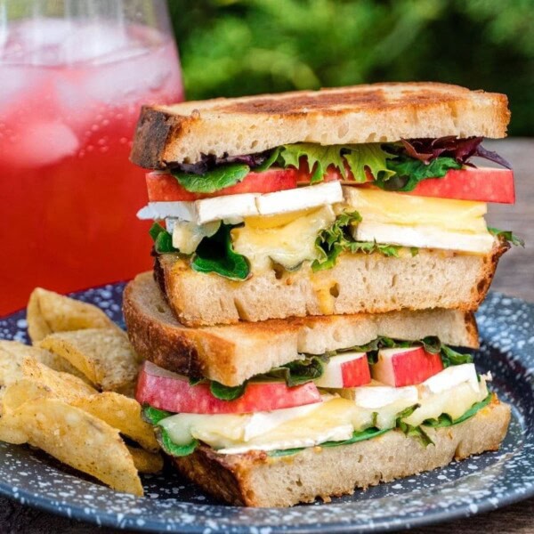 A plate with a sandwich containing lettuce, tomato, and cheese, potato wedges, and a glass of iced pink drink in the background.