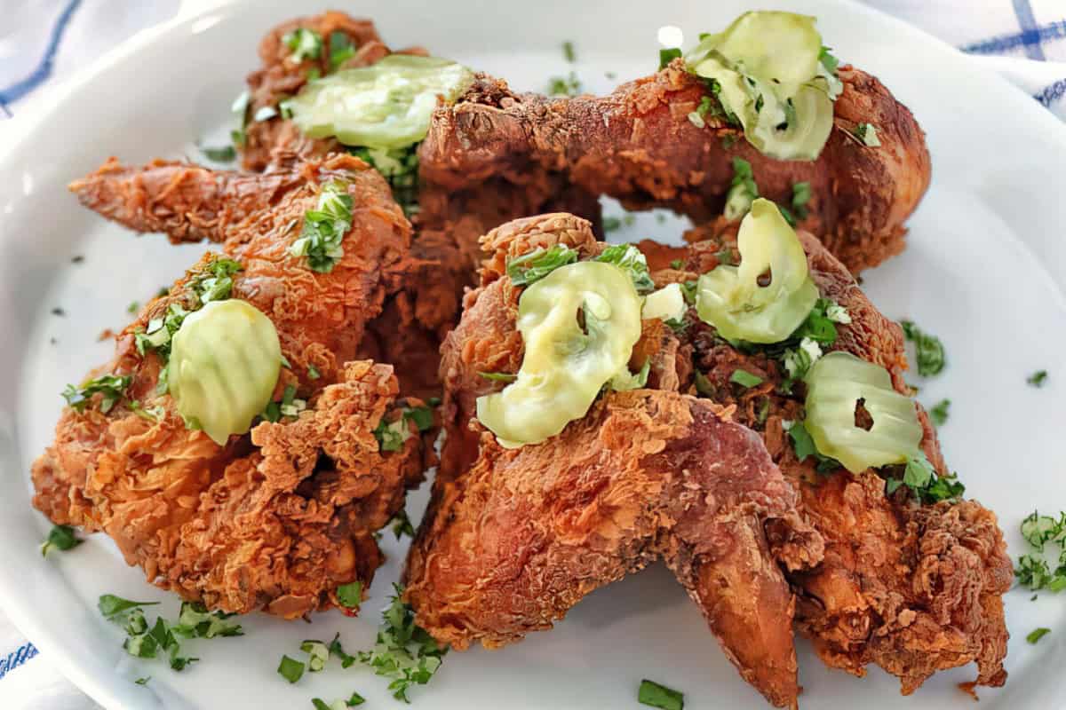 Plate of crispy fried chicken wings topped with sliced pickles and sprinkled with chopped herbs, arranged on a white dish.