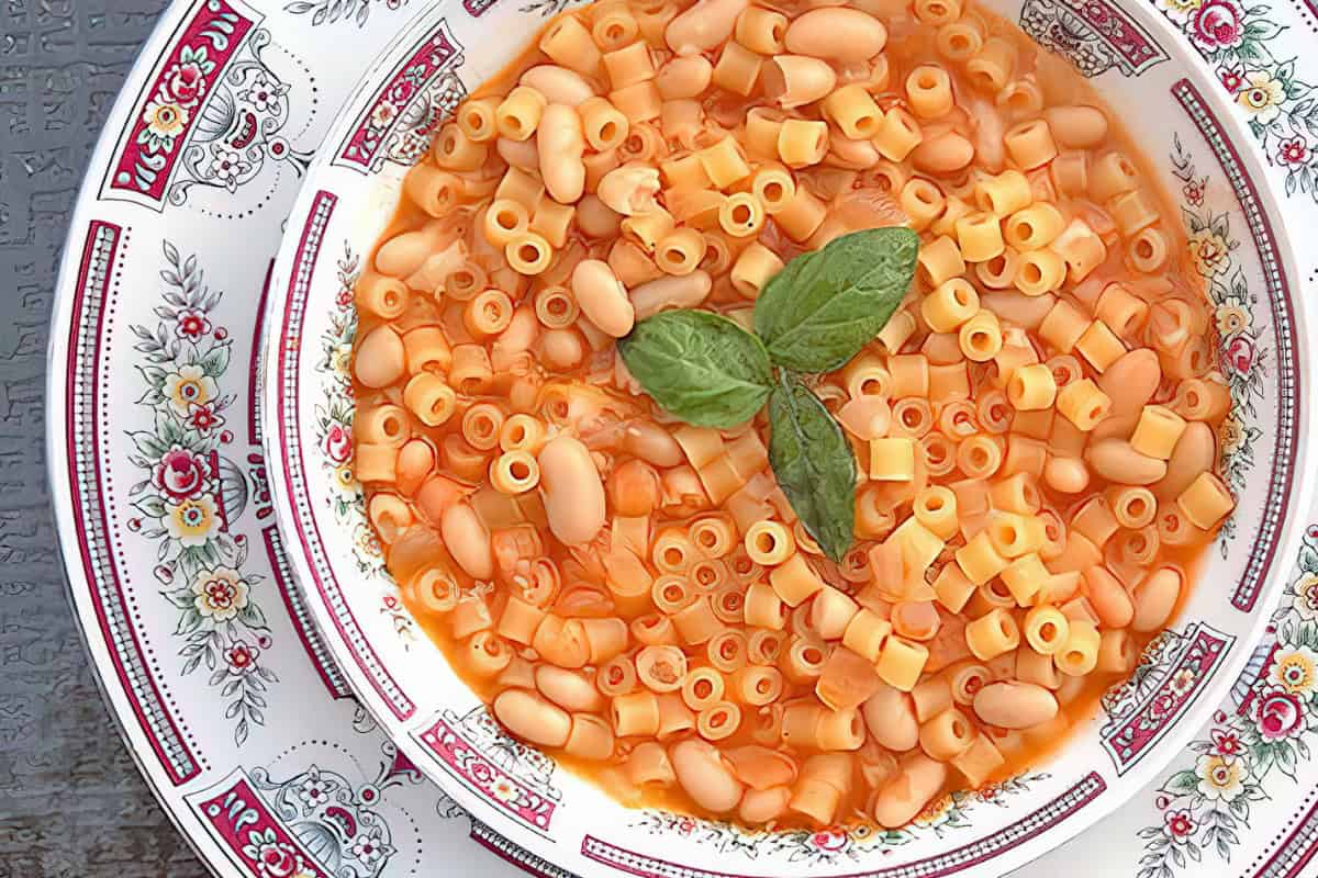 A bowl of pasta e fagioli containing small pasta and beans in tomato broth, garnished with basil, on a decorative plate.