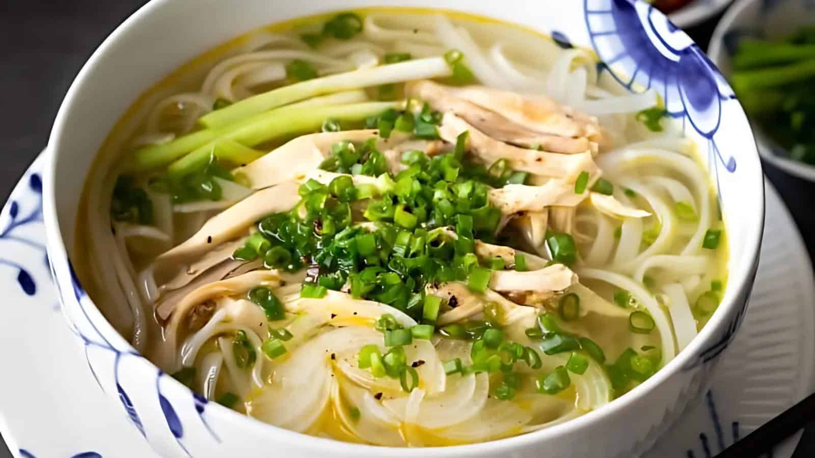 A bowl of Vietnamese pho featuring rice noodles, sliced chicken, and chopped green onions in a clear broth. The bowl has a decorative blue and white pattern on the rim.