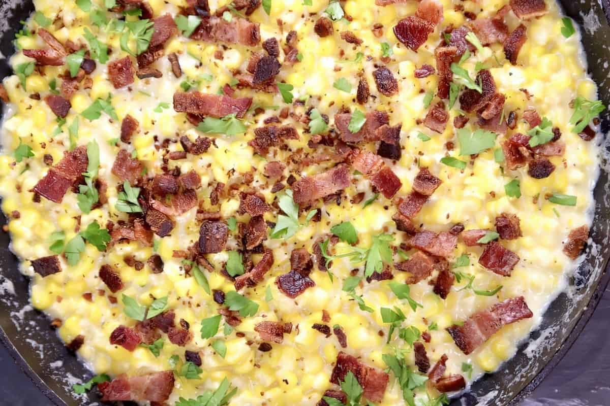 A skillet containing creamy corn garnished with crispy bacon pieces and chopped parsley.