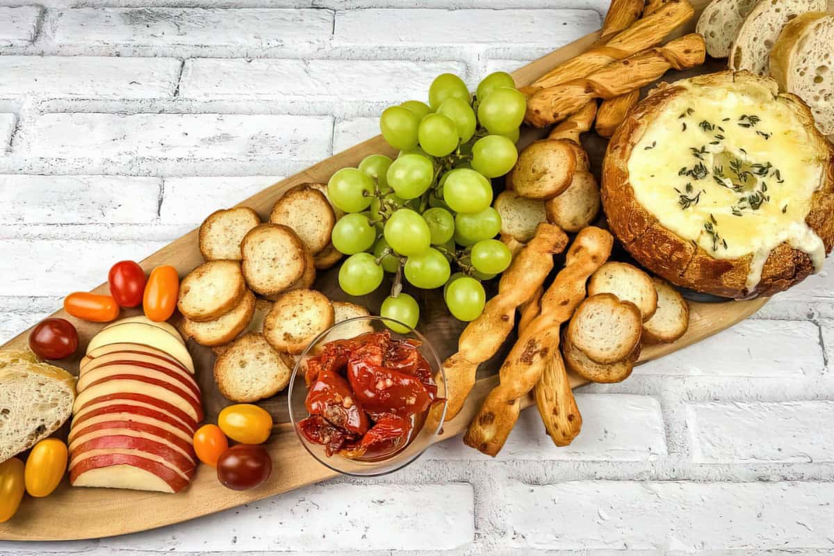A charcuterie board with bread, green grapes, cherry tomatoes, sliced apples, breadsticks, and a bread bowl with melted cheese. A small dish holds sun-dried tomatoes. The board rests on a light brick background.