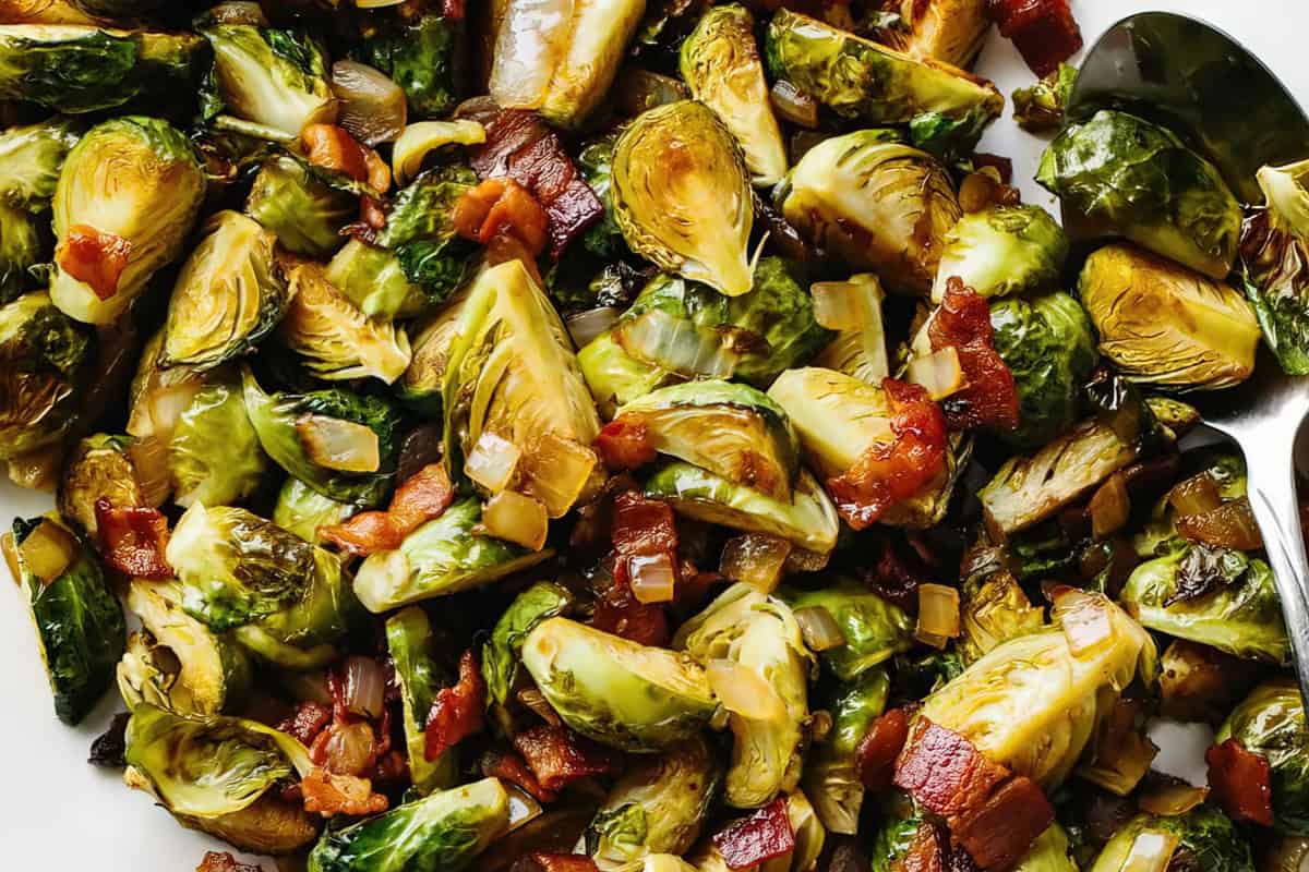 A close-up of roasted Brussels sprouts mixed with crispy bacon and onions on a white surface. The dish appears savory and well-cooked, with golden brown and green tones, accompanied by a silver serving spoon.