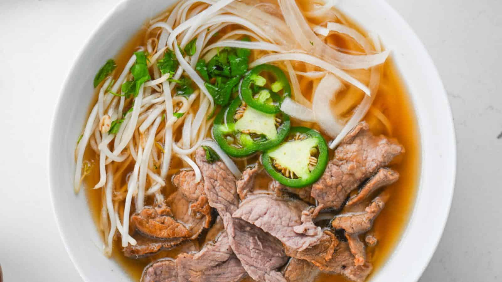A bowl of pho with thinly sliced beef, rice noodles, bean sprouts, sliced green jalapeños, and chopped green herbs in a clear broth.