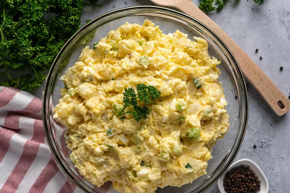 A glass bowl of potato salad garnished with parsley, surrounded by a striped cloth, a wooden spatula, and scattered peppercorns.