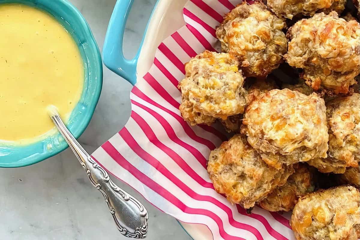 A plate of sausage balls is on pink striped paper, accompanied by a blue bowl containing yellow dipping sauce with a spoon.