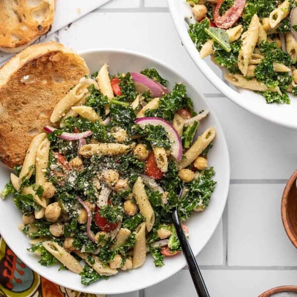 A bowl of penne pasta salad with kale, chickpeas, red onions, tomatoes, and grated cheese beside toasted bread on white tiles.