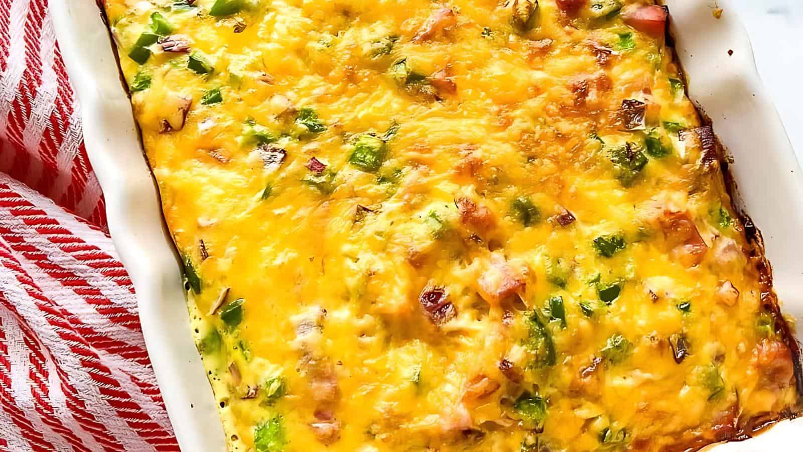 A baked casserole in a white dish featuring melted cheese, green peppers, and bits of meat is positioned next to a red and white striped cloth.