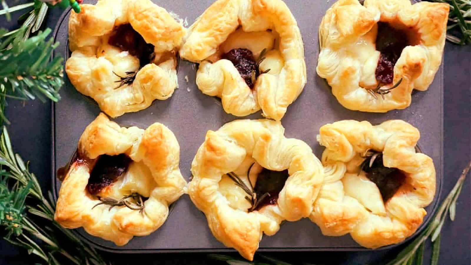 Six puff pastry appetizers filled with melted cheese and a dark filling, garnished with rosemary, are arranged in a baking tray. Flaky and golden, the pastries are surrounded by evergreen branches.