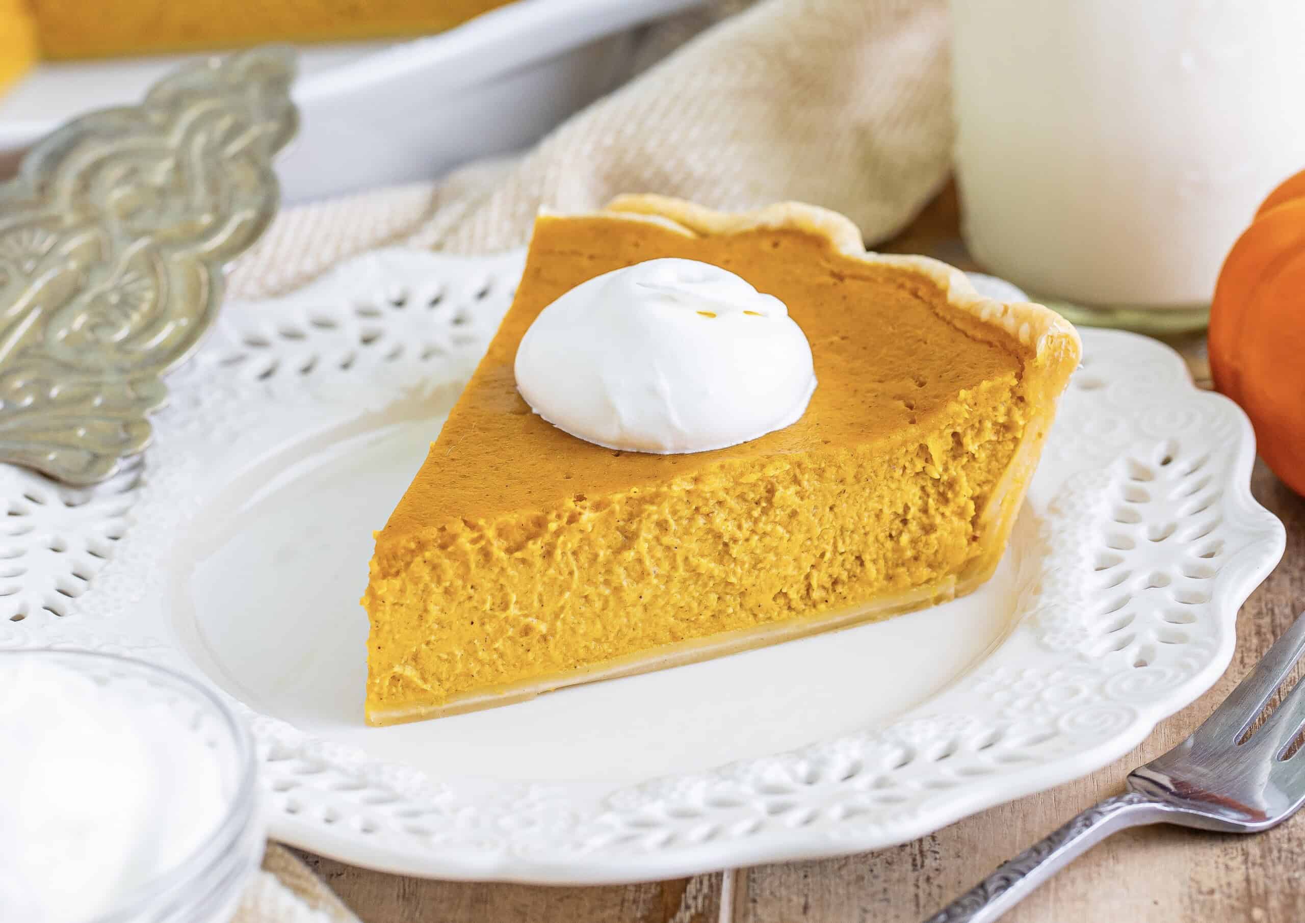 The image features a slice of pumpkin pie topped with whipped cream, placed on a white plate. Nearby are a fork and a glass of milk.