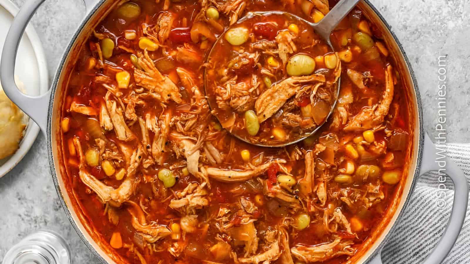 The image shows a pot of chicken stew containing corn, lima beans, and a tomato-based broth. A ladle is shown scooping some of the stew.