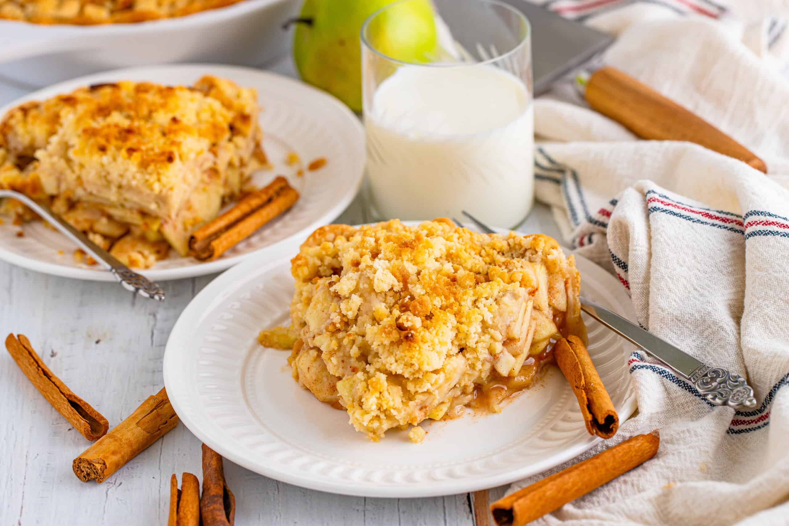 The image depicts apple crumble dessert served on plates, accompanied by cinnamon sticks, a glass of milk, and a striped cloth, all set on a white wooden table.