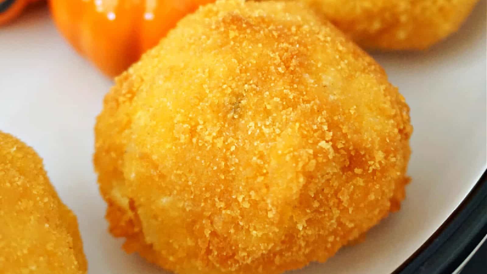 A close-up of a golden, crispy fried ball, possibly a croquette or arancini, on a white plate. In the background, there is a hint of an orange garnish, possibly a small pepper. The food appears appetizing and crunchy.