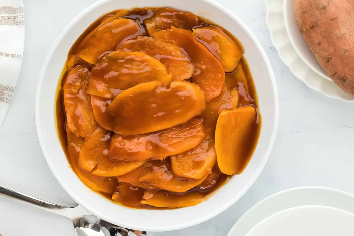 A white bowl containing candied sweet potatoes in thick syrup is situated on a light-colored surface, accompanied by a spoon and a plate.