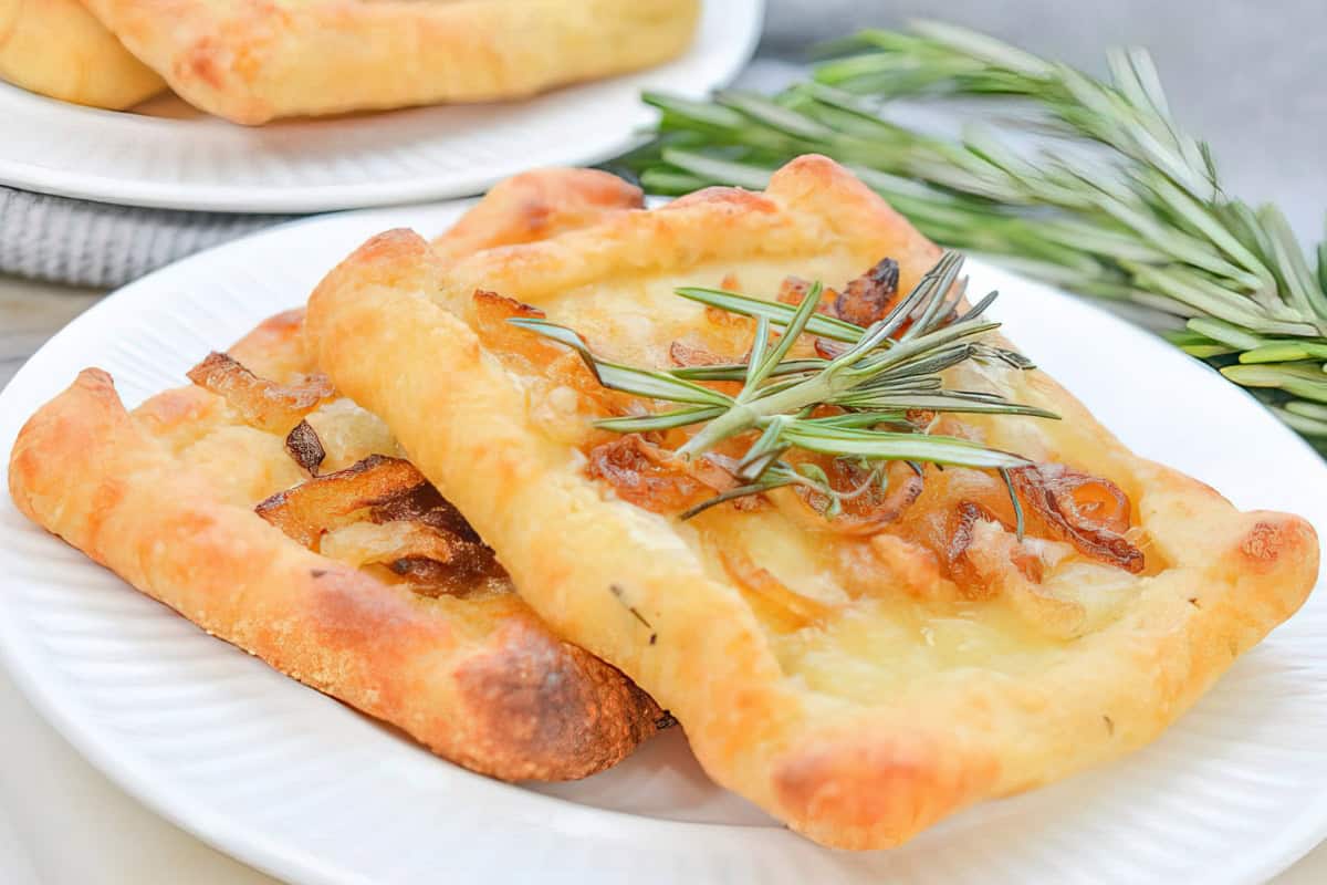 Golden rectangular pastries topped with caramelized onions and sprigs of rosemary are placed on a white plate. Another similar plate is partly visible in the background. Fresh rosemary lies beside the plate.