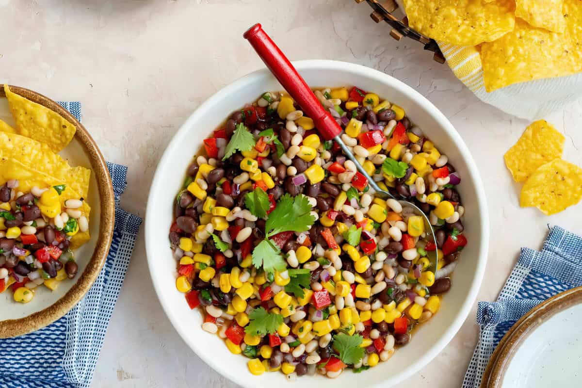 A bowl of corn and bean salad with red bell peppers, onions, and cilantro is placed next to a small dish of tortilla chips and a larger basket filled with more chips on a textured surface.