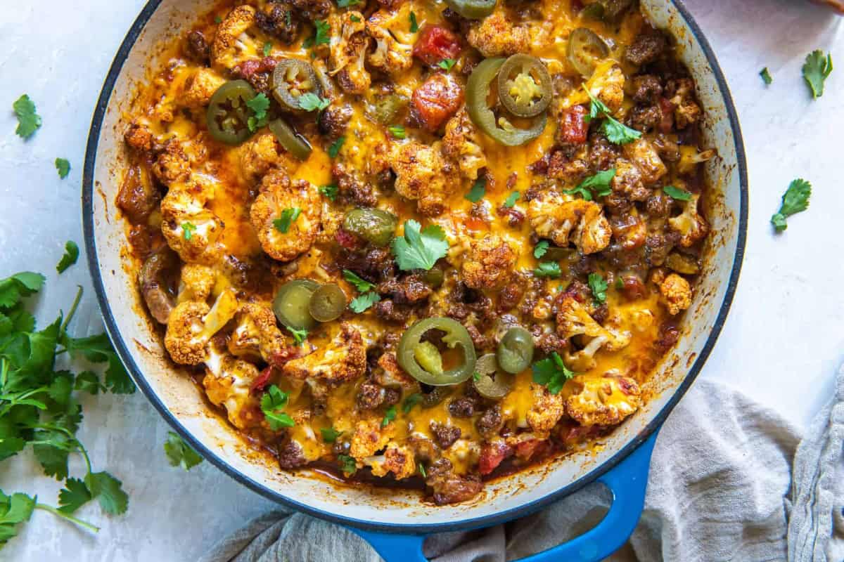The image shows a skillet with melted cheese, ground meat, cauliflower, sliced jalapeños, and chopped cilantro.
