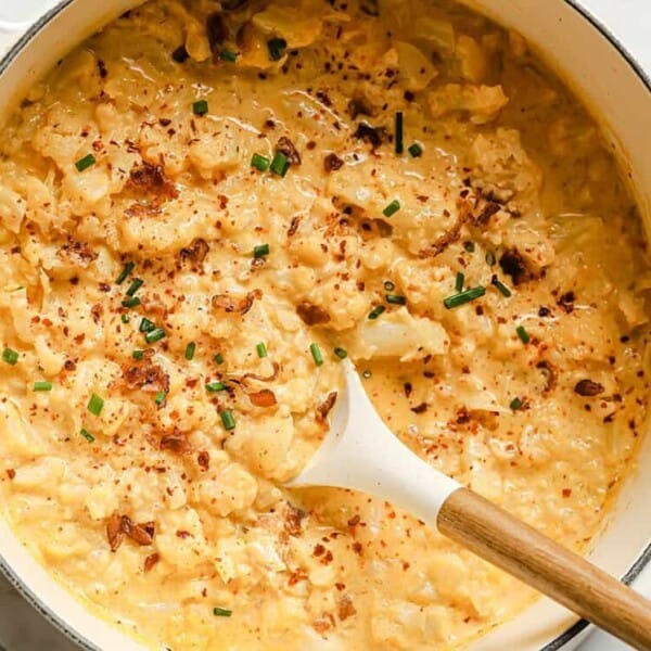 A pot of creamy scrambled eggs topped with chopped chives and red pepper flakes, with a wooden spoon resting inside.