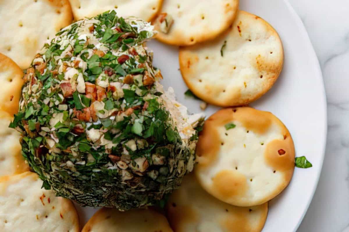 A cheese ball covered in chopped nuts and herbs is placed on a white plate surrounded by round crackers. The dish is set on a marble countertop, offering a savory appetizer presentation.