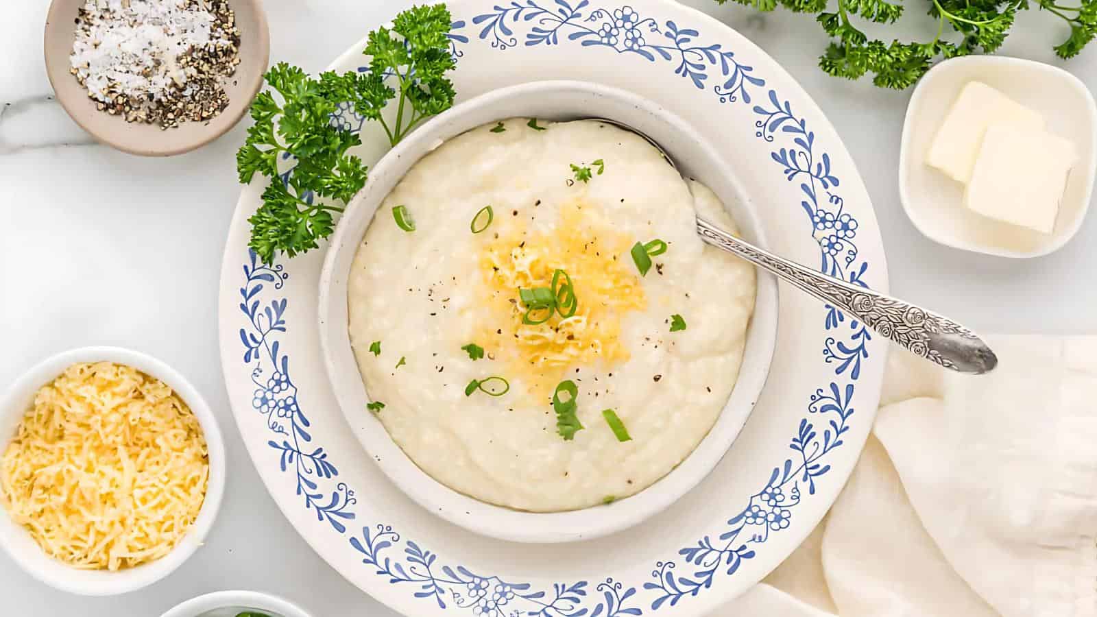 The image depicts a bowl of grits topped with cheese, green onions, and garnished with parsley. Nearby are small dishes containing shredded cheese, butter, and salt and pepper.