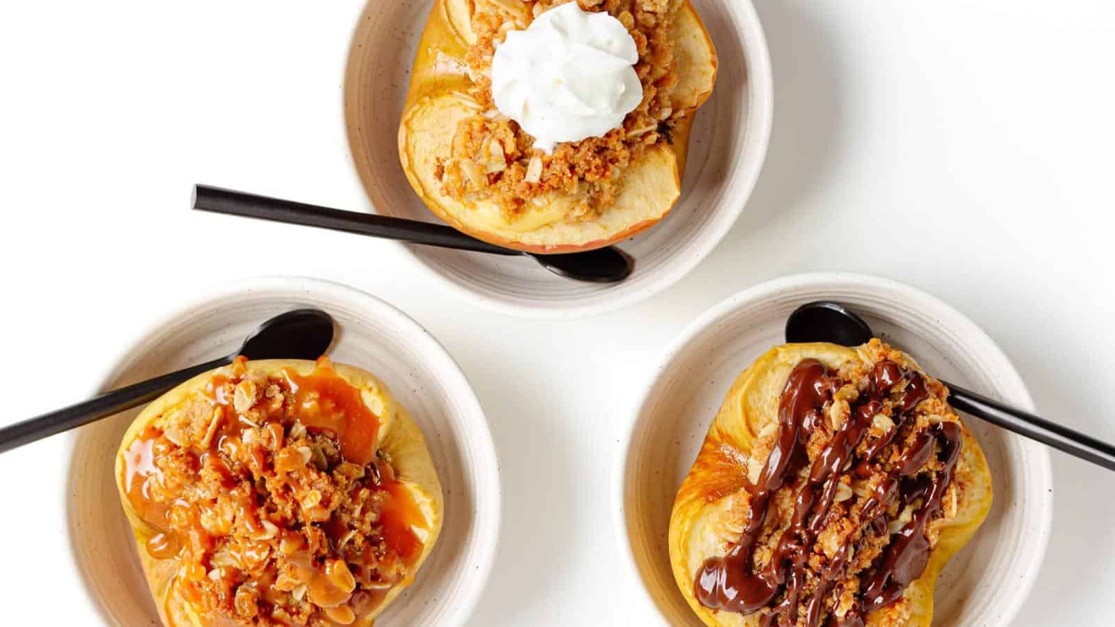 Three bowls, each with a baked apple topped with different toppings: whipped cream, caramel sauce with nuts, and chocolate sauce with nuts. Each bowl has a black spoon on a white background.