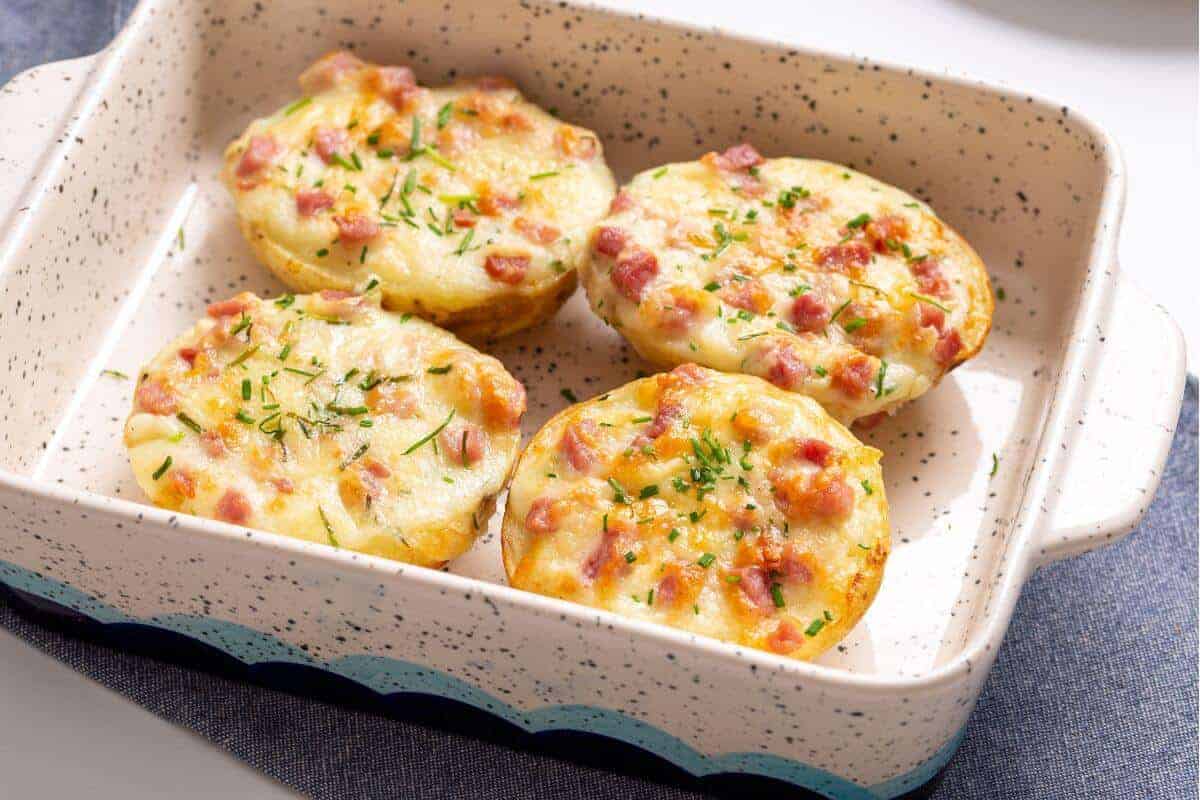 The image shows four mini potato casseroles topped with cheese, diced ham, and herbs in a speckled ceramic baking dish.
