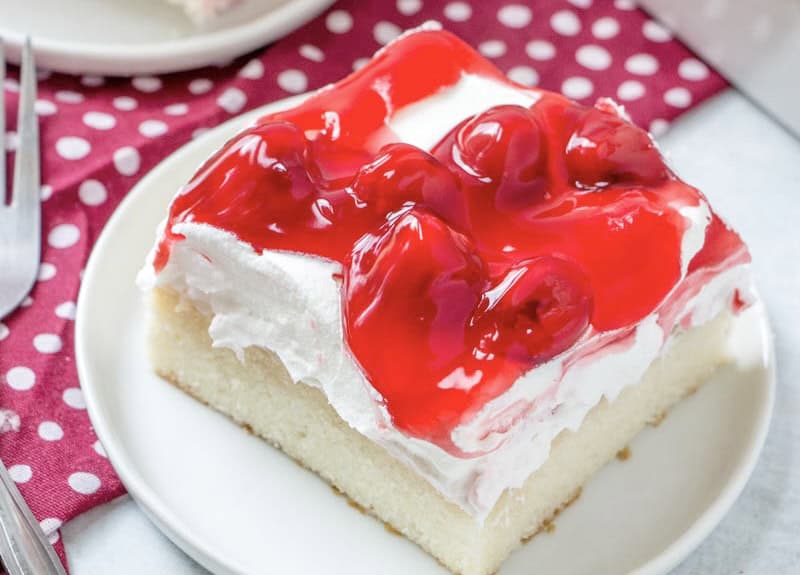 A slice of cake with white frosting and cherry topping on a white plate, accompanied by a red polka dot napkin.