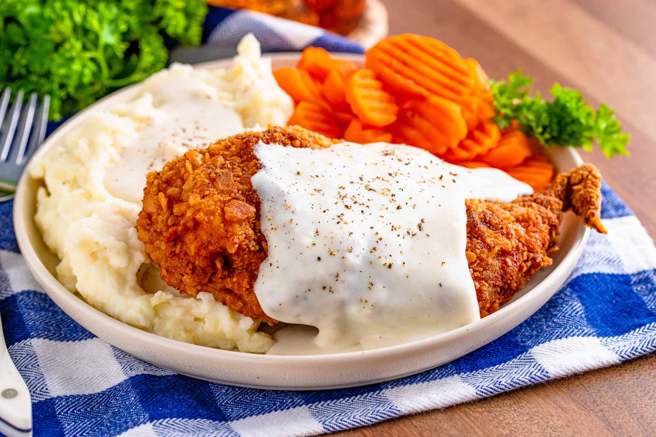 A plate of fried chicken covered in gravy, accompanied by mashed potatoes and sliced carrots, set on a checkered cloth.