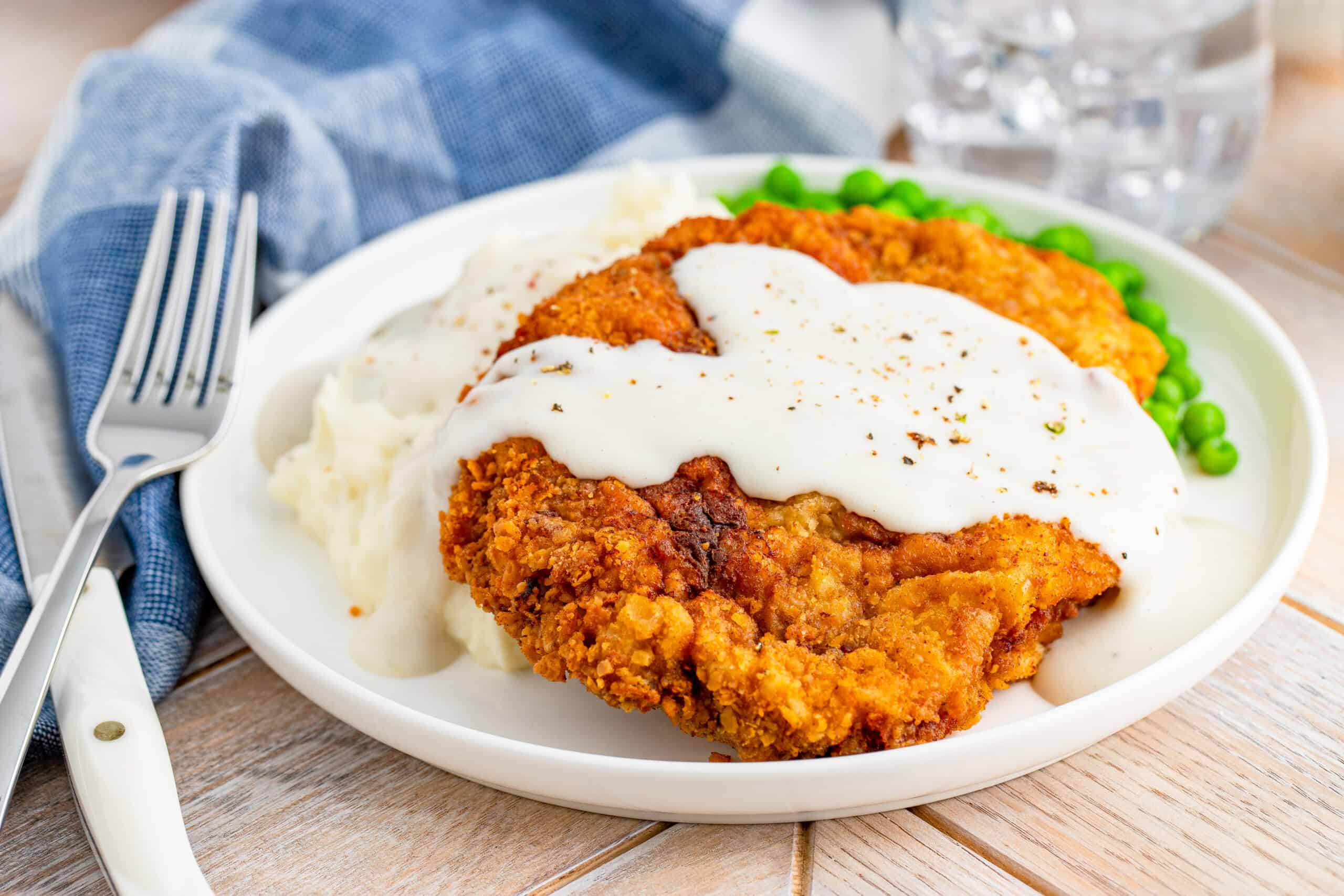 The image depicts a plate containing fried chicken with white gravy, mashed potatoes, and green peas. Beside the plate are a fork and a blue napkin.