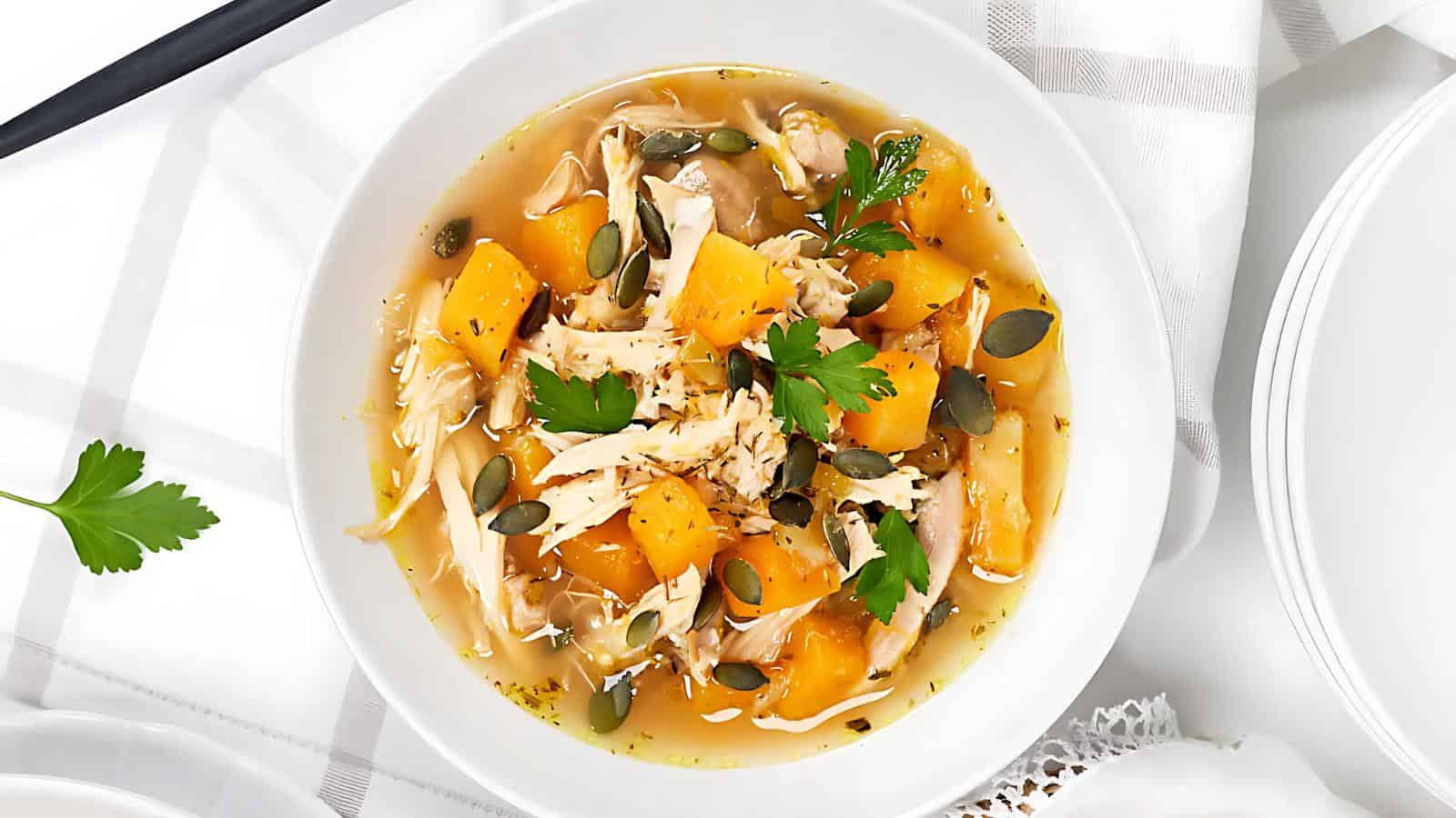 A bowl of chicken soup with chunks of butternut squash, garnished with parsley and pumpkin seeds, on a white cloth. The soup is in a white bowl, providing a comforting and hearty appearance.