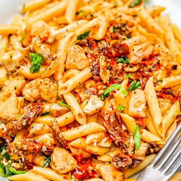 A plate of penne pasta with shrimp, sun-dried tomatoes, herbs, and sauce, garnished with greens and a fork on the side.
