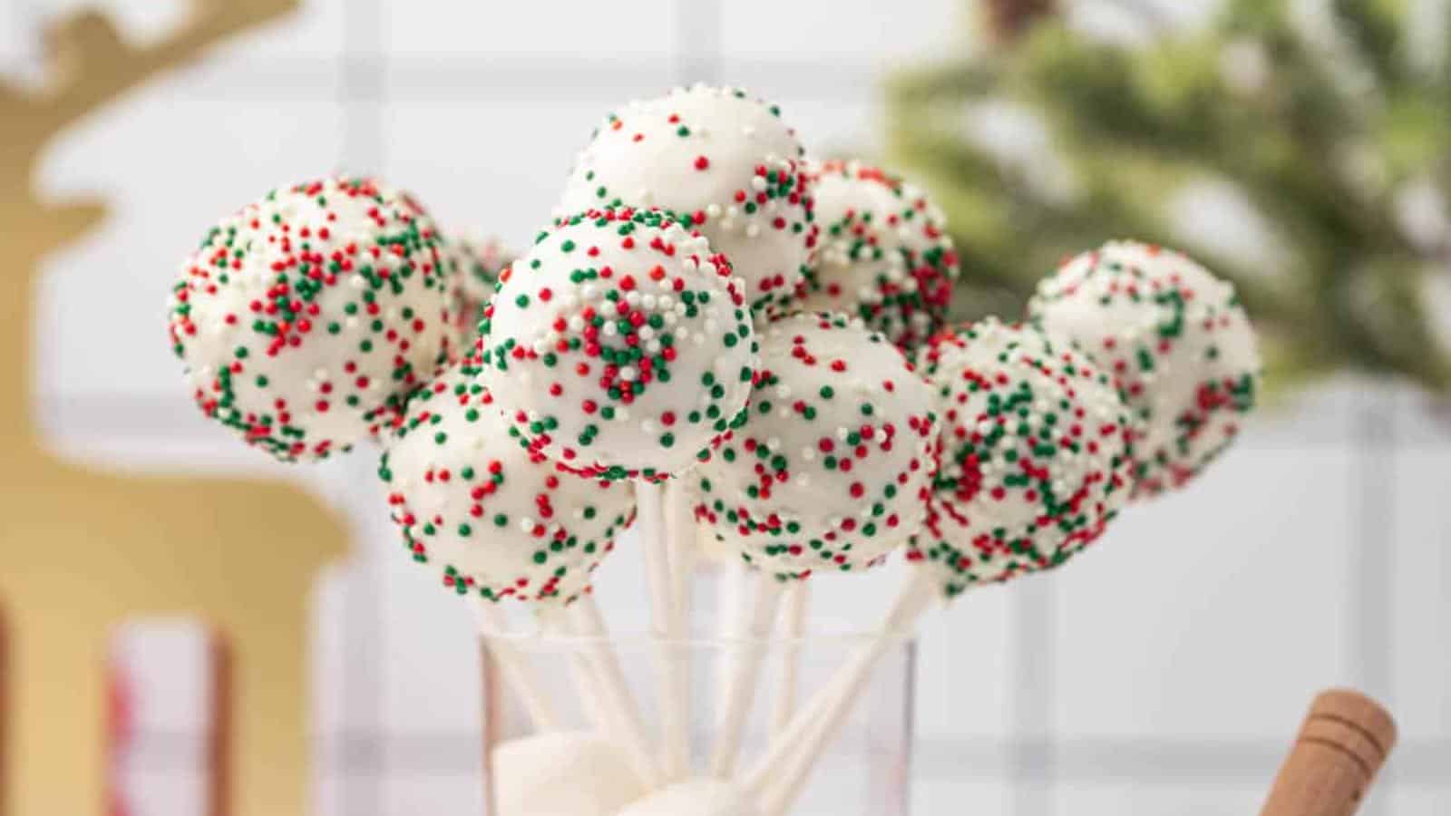 A glass holds several cake pops coated in white chocolate with red, green, and white sprinkles. They are arranged like a bouquet. In the background, blurred Christmas decorations are visible.