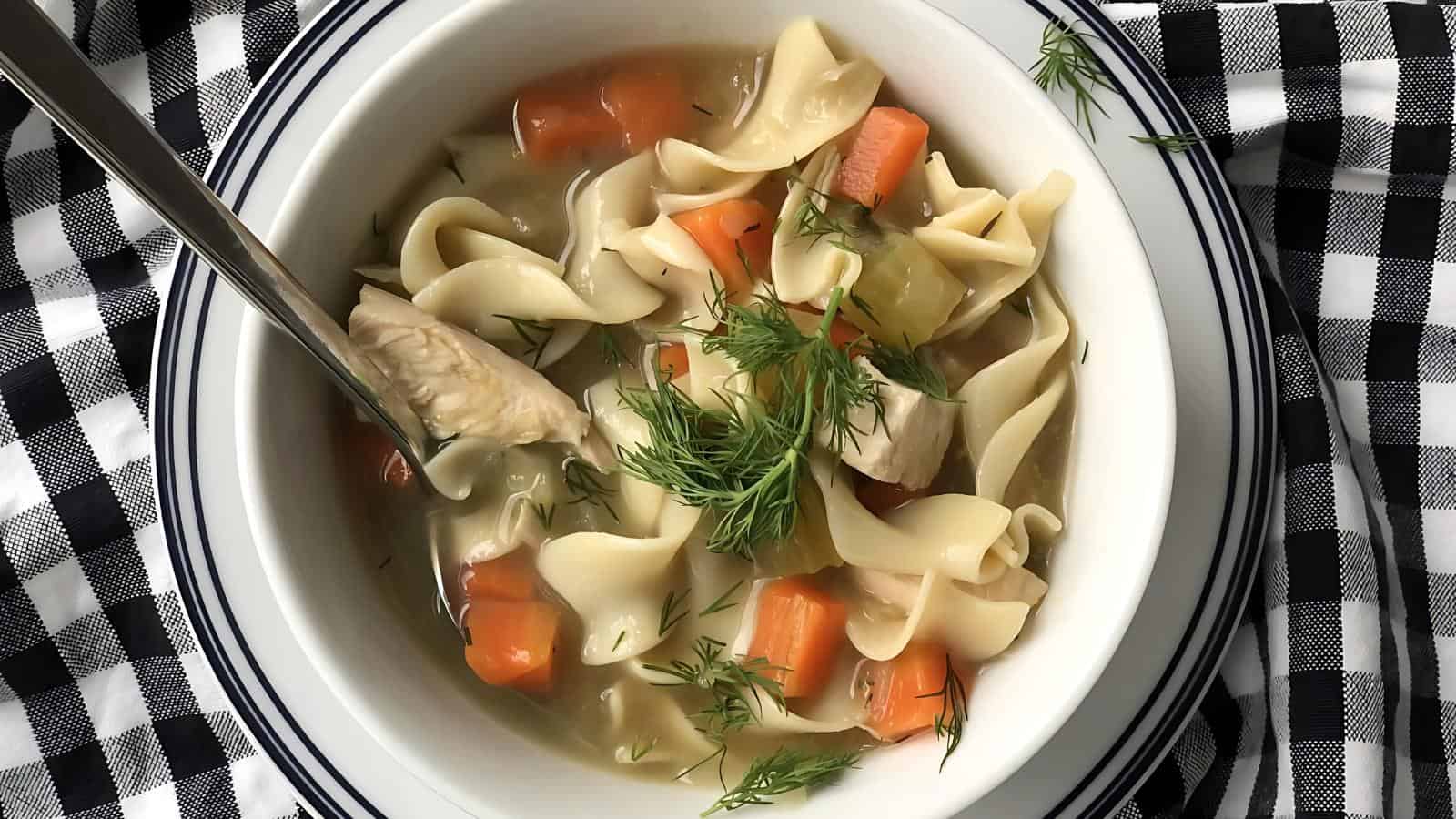 A bowl of chicken noodle soup with carrots and herbs on a checkered cloth.