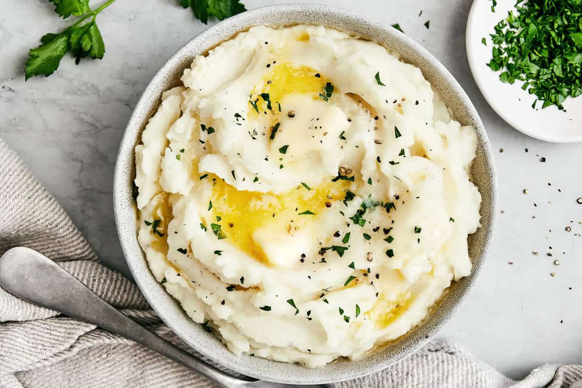 A bowl of creamy mashed potatoes topped with melted butter and sprinkled with chopped parsley. A fork rests beside the bowl on a light cloth, and there are parsley leaves and a small bowl of more chopped parsley nearby.