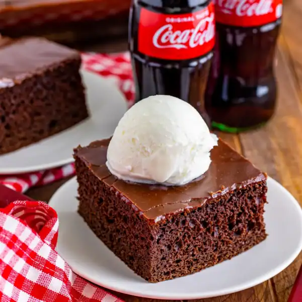 A slice of chocolate cake with vanilla ice cream on a white plate, beside a red checkered napkin and two bottles of Coca-Cola.